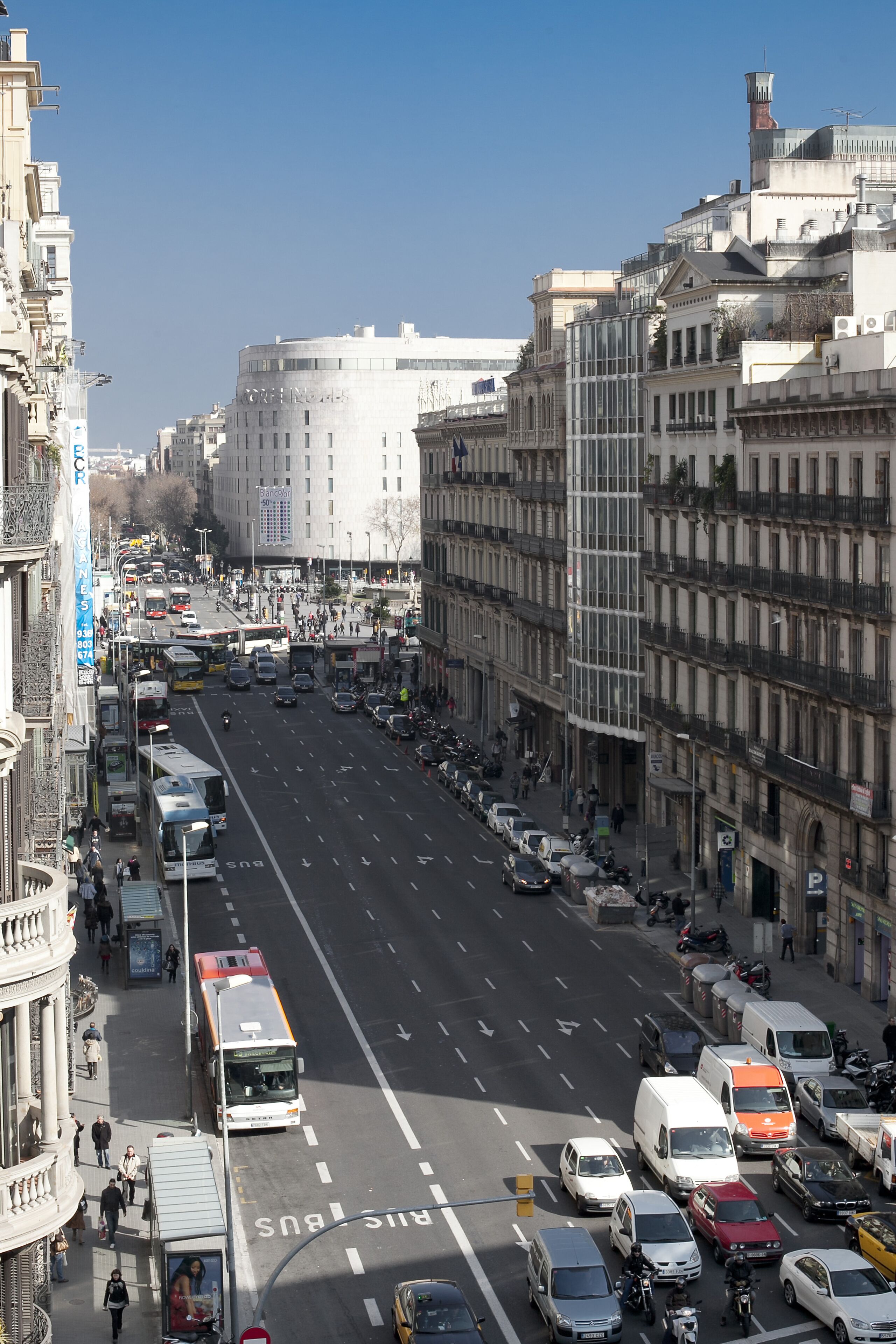 A busy urban street with a mix of modern and historic buildings, a clear sky, and various vehicles including buses and cars.