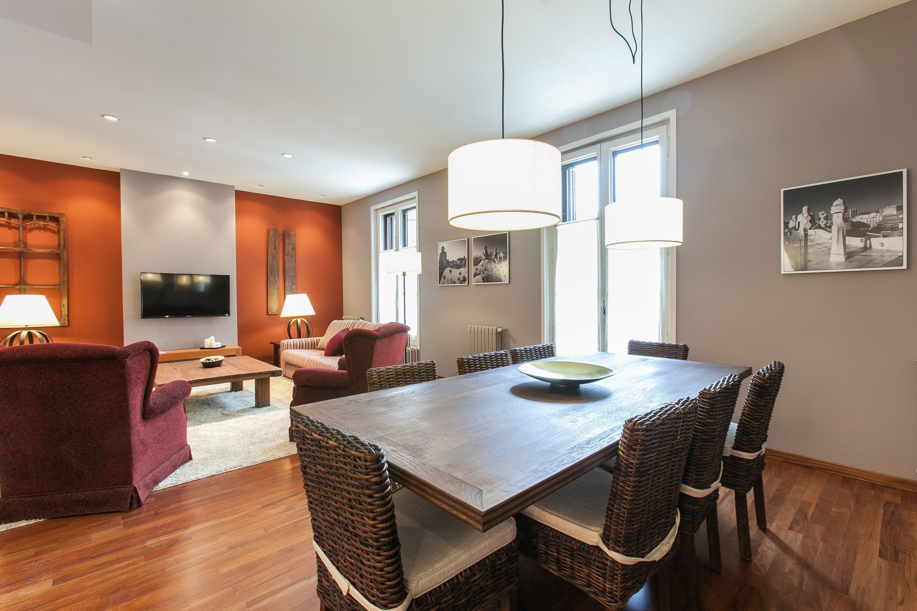 A modern living room with a wooden dining table, wicker chairs, a flat-screen TV, and a large window providing natural light.