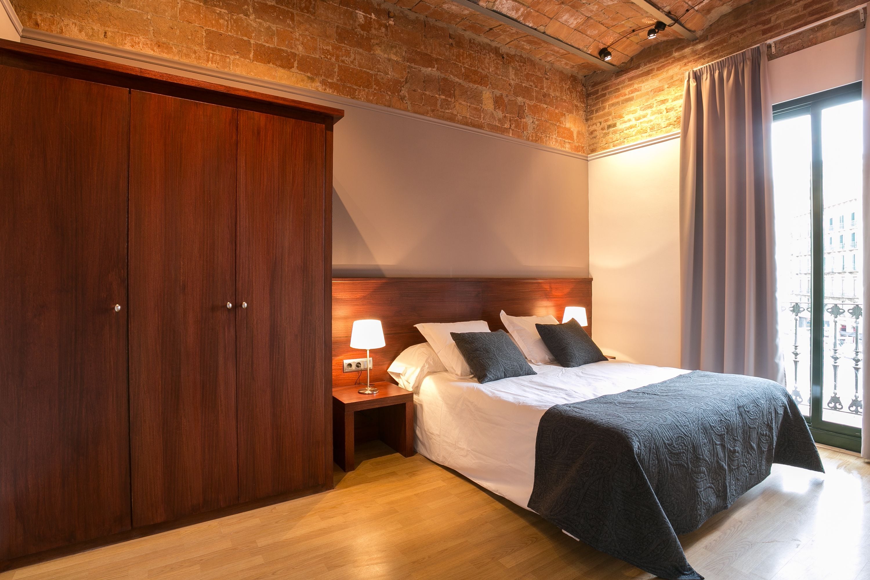 A bedroom with a large wardrobe, a bed with a dark bedspread, a nightstand with a lamp, and a view of the outside through a window with curtains.