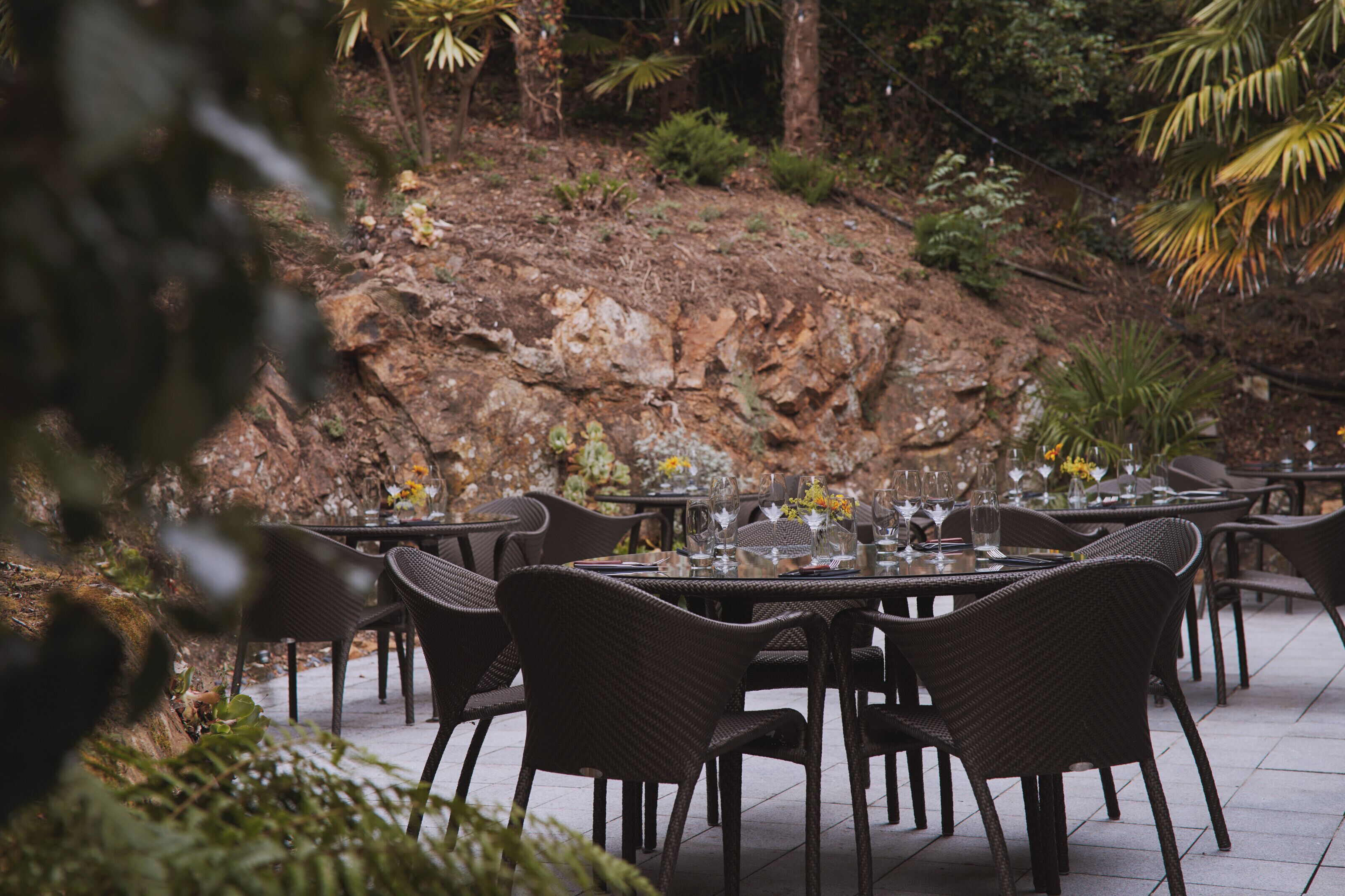An outdoor dining area with wicker chairs and tables, surrounded by greenery and a rocky hillside.