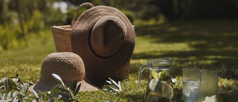 A straw hat, a woven basket, a glass pitcher with mint, and two clear glasses on a grassy field.
