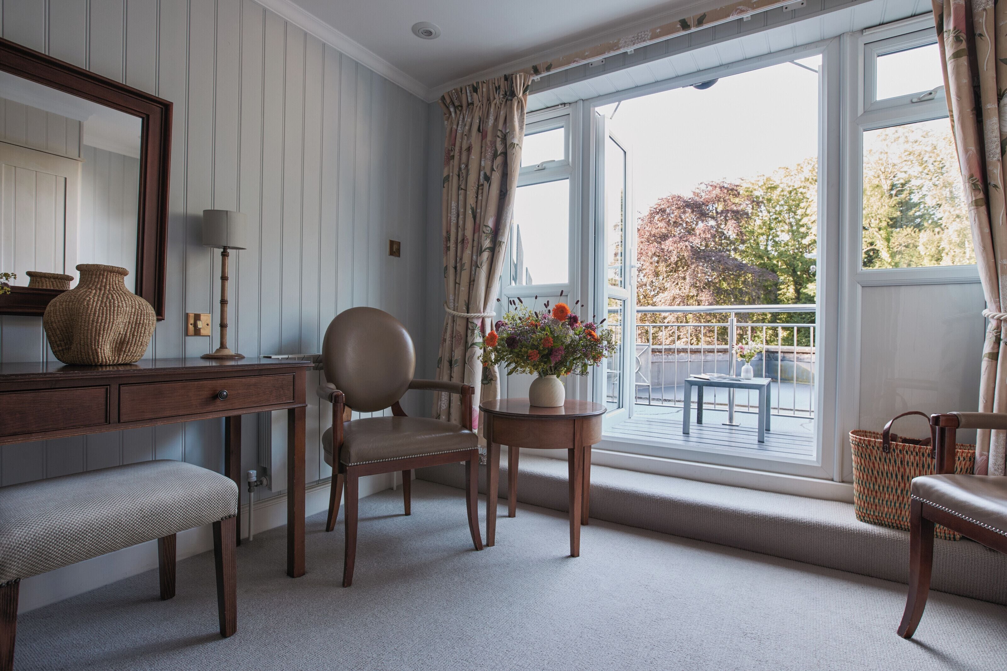 A room with a wooden desk, a chair, a bench, a table with a vase of flowers, and a view of the outdoors through a large window.