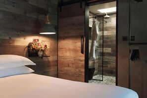 A modern bedroom with a wooden wall, a bed with white linens, a bedside table with a vase of flowers, and a glass shower enclosure. - Fermain Valley Hotel (St. Peter Port)