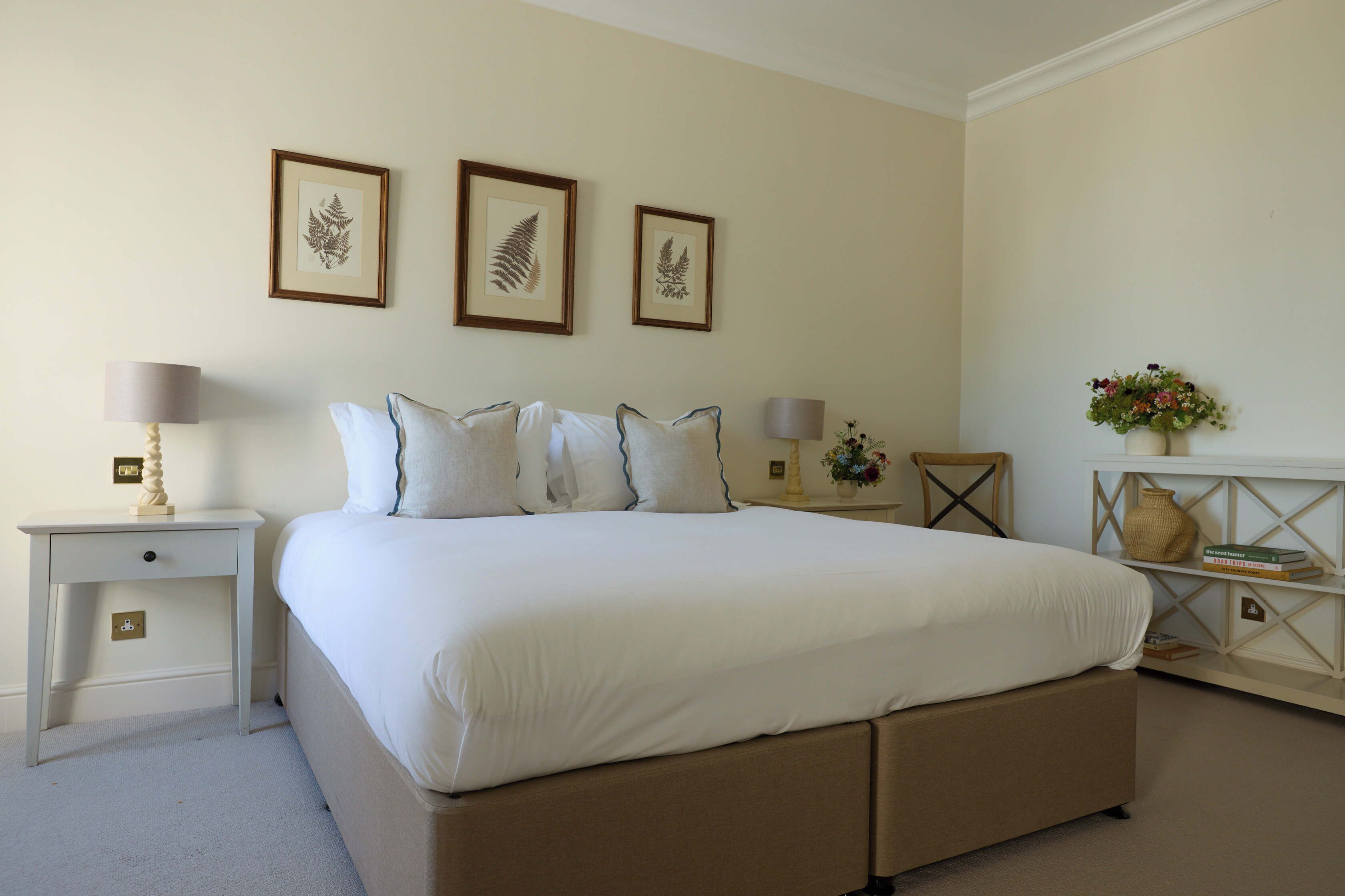A bedroom with a bed, nightstands, a lamp, framed artwork, and a vase with flowers.