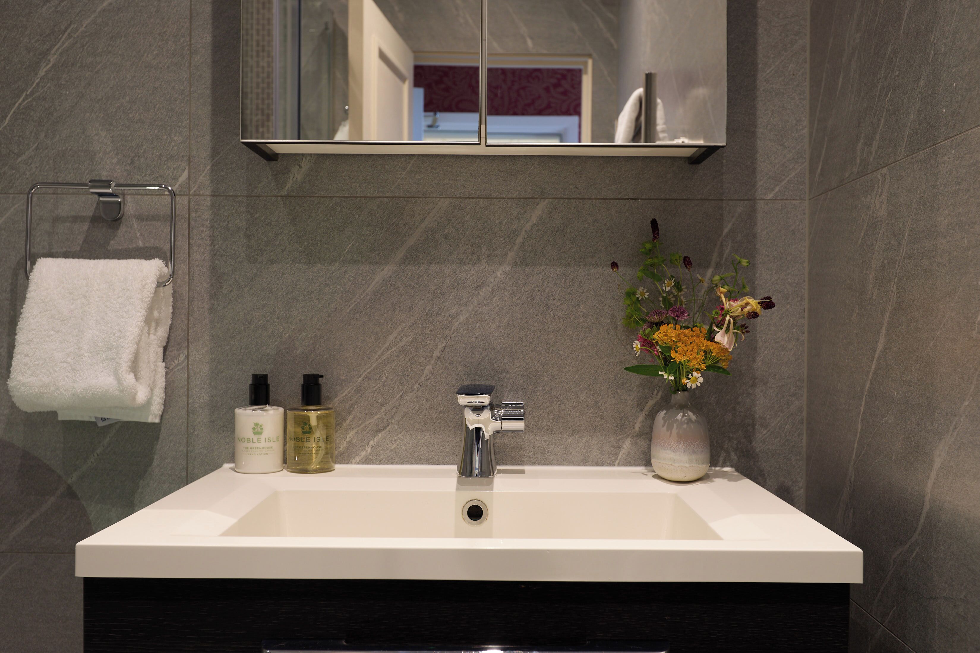 A bathroom with a white sink, a mirror above it, a towel rack with a white towel, and a vase with flowers.
