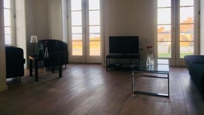 Una sala de estar con televisor, sofá, mesa de centro y una silla. Amplias ventanas ofrecen vistas al exterior.
