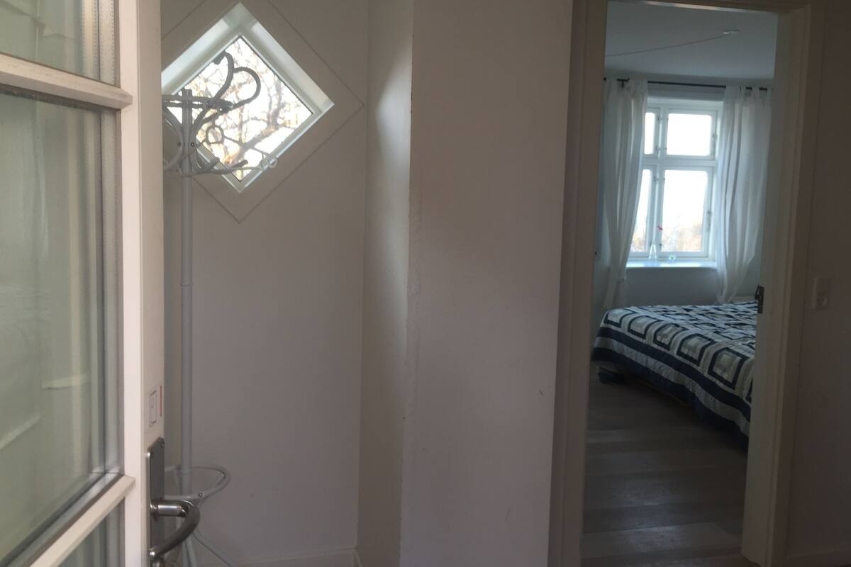 Ein Flur, der zu einem Schlafzimmer führt, mit einem Bett, einem Fenster mit Vorhängen und einer Tür mit Griff.