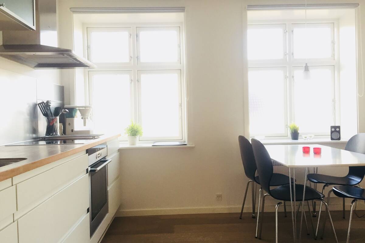 Eine moderne Küche mit weißen Schränken, einer Edelstahl-Dunstabzugshaube und einem Fenster mit Blick. Ein Esstisch mit schwarzen Stühlen und ein kleiner Topfpflanze stehen auf der Fensterbank.