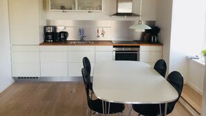 Una cocina moderna con armarios blancos, una campana extractora de acero inoxidable, horno integrado y un comedor con mesa blanca y sillas negras.