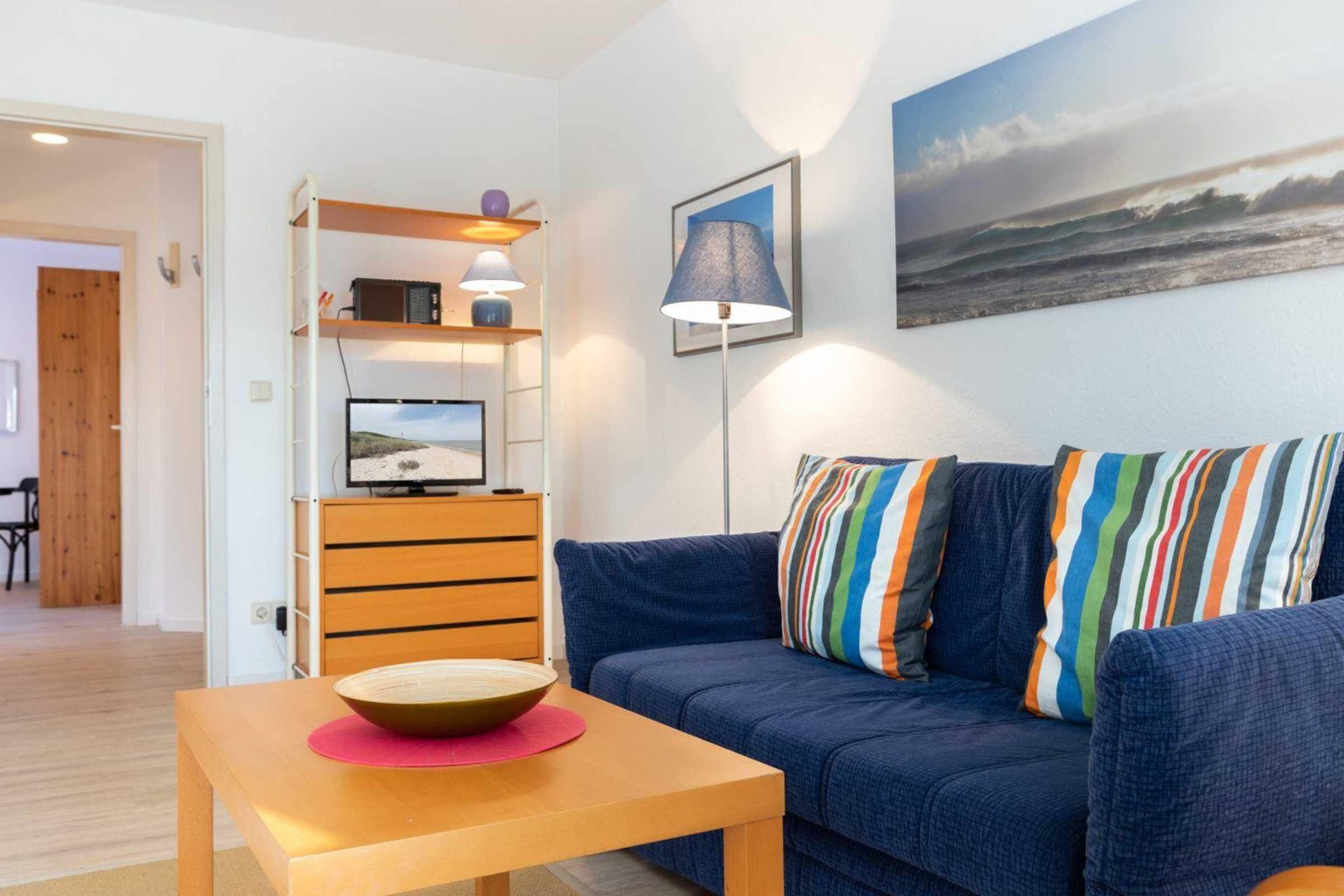 A modern living room with a blue sofa, colorful striped pillows, a wooden coffee table, a flat-screen TV on a wooden stand, and a framed landscape painting on the wall.