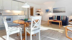 A modern kitchen with white cabinets, a wooden dining table, and a blue sofa in a living area. - Apartment 1 (ID 088), Germany (Sankt Peter-Ording)