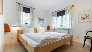 A bedroom with a wooden bed, two bedside tables, a chair, and a window with curtains. - Apartment 1 (ID 088), Germany (Sankt Peter-Ording)