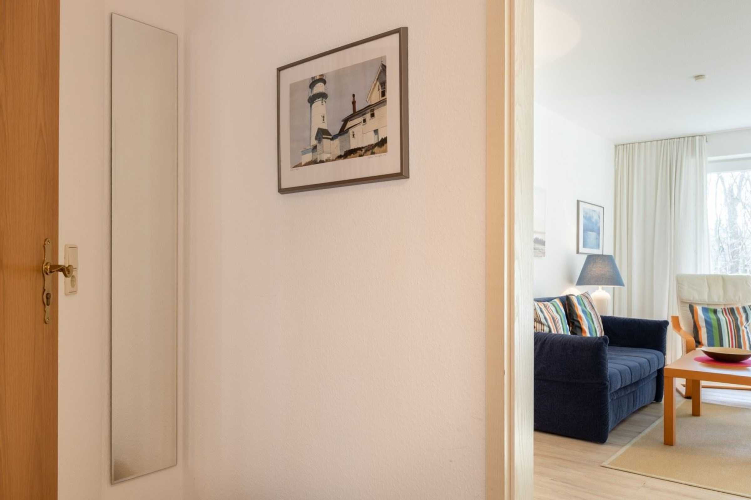 A room with a large mirror, a framed picture of a lighthouse, a dark blue sofa, a wooden table, and a lamp.
