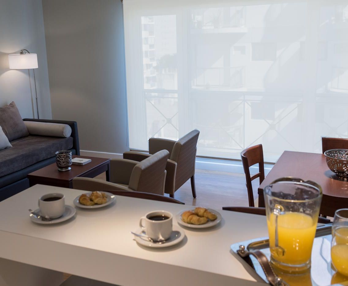 A modern hotel room with a dining area, a sofa, and a balcony with a view of buildings.