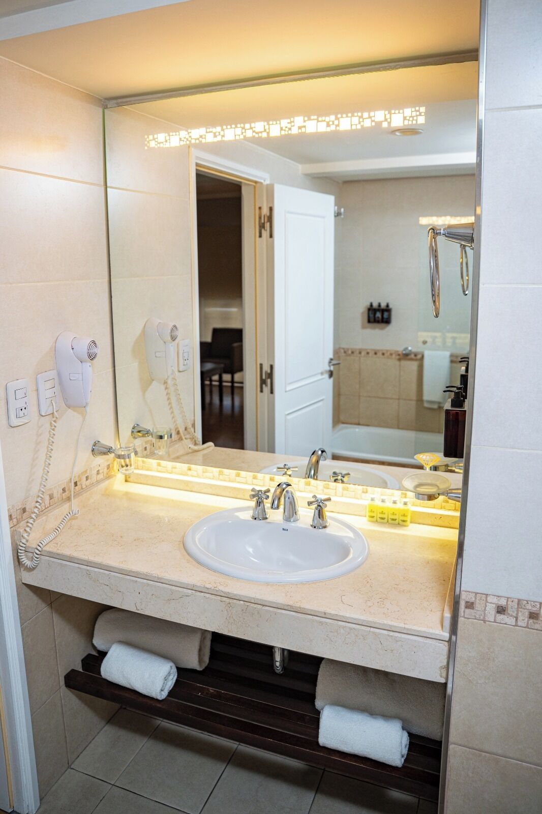 Un baño con un lavamanos blanco, dos grifos, un espejo, un secador de pelo y un toallero.
