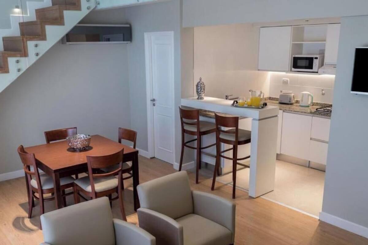 A modern kitchen with white cabinets, a microwave, and a coffee maker. There is a dining area with a wooden table and chairs.