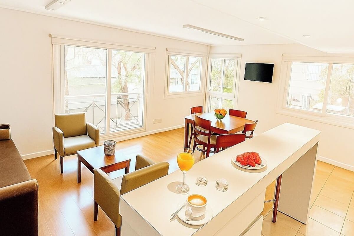 A bright, modern kitchen with a central island, dining area, and large windows.