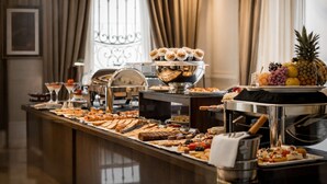 A buffet with various food items, including pastries, fruits, and desserts, arranged on a counter with a coffee machine and a decorative metal railing in the background. - Carat Apart Boutique (Rosario)