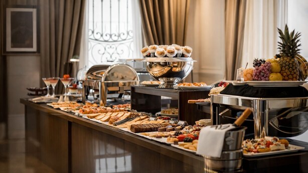 A buffet with various food items, including pastries, fruits, and desserts, arranged on a counter with a coffee machine and a decorative metal railing in the background. - Carat Apart Boutique (Rosario)