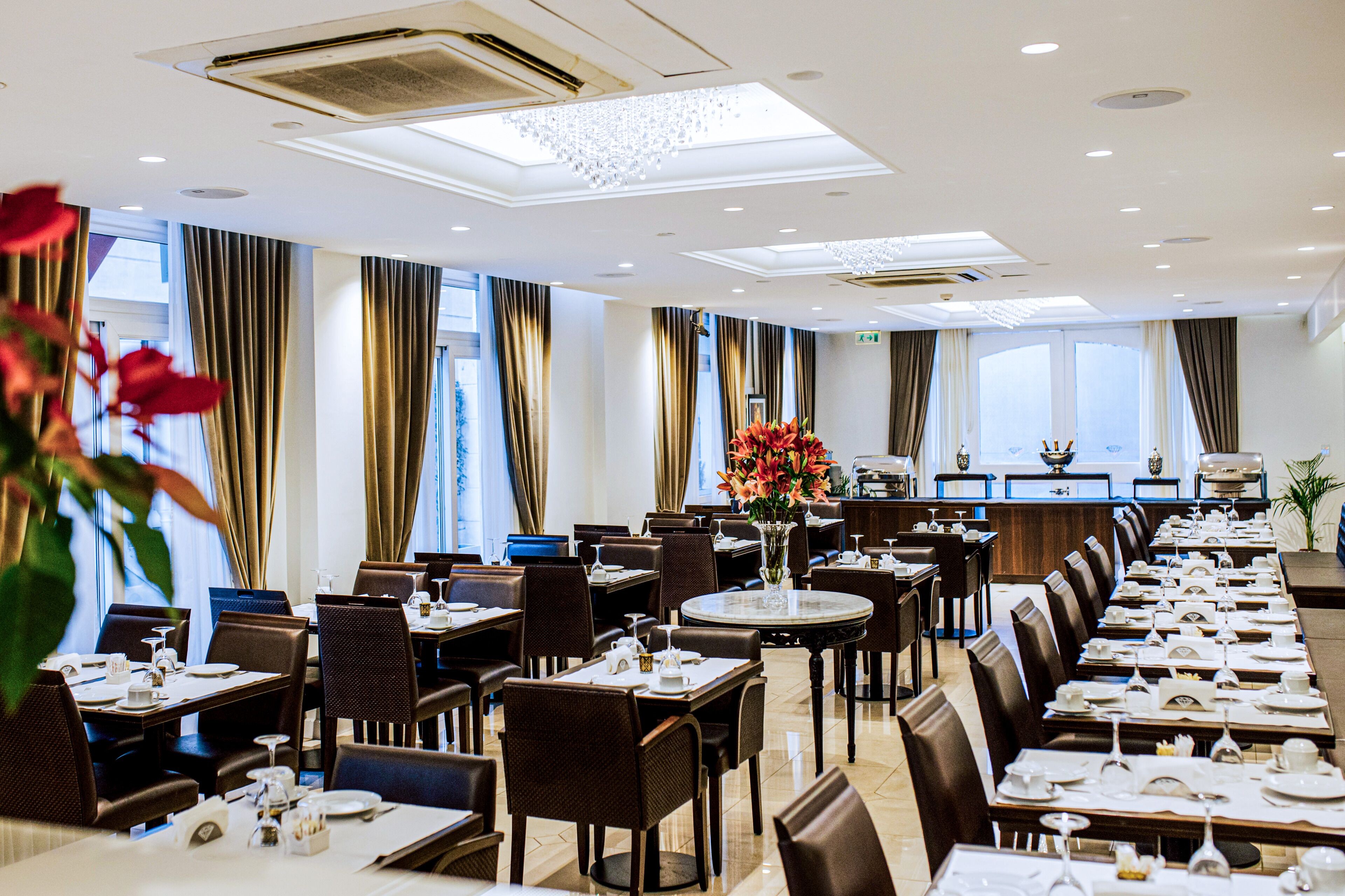 Un comedor de hotel con varias mesas y sillas, un arreglo floral central y vista a la cocina al fondo.
