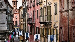 A narrow street lined with colorful buildings, some with balconies and shutters, and a clock tower in the background. - Le Camere di Gio (Alba)