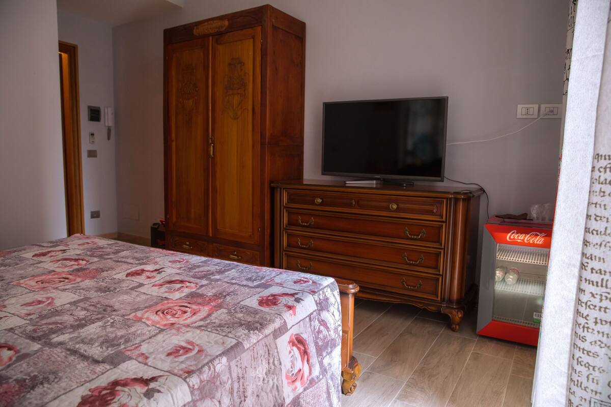 A bedroom with a bed, a wooden wardrobe, a television on a dresser, and a refrigerator.