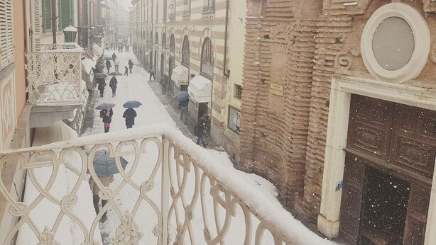 A snow-covered street with buildings, a balcony with ornate metal railings, and a brick wall with a circular window. - Le Camere di Gio (Alba)