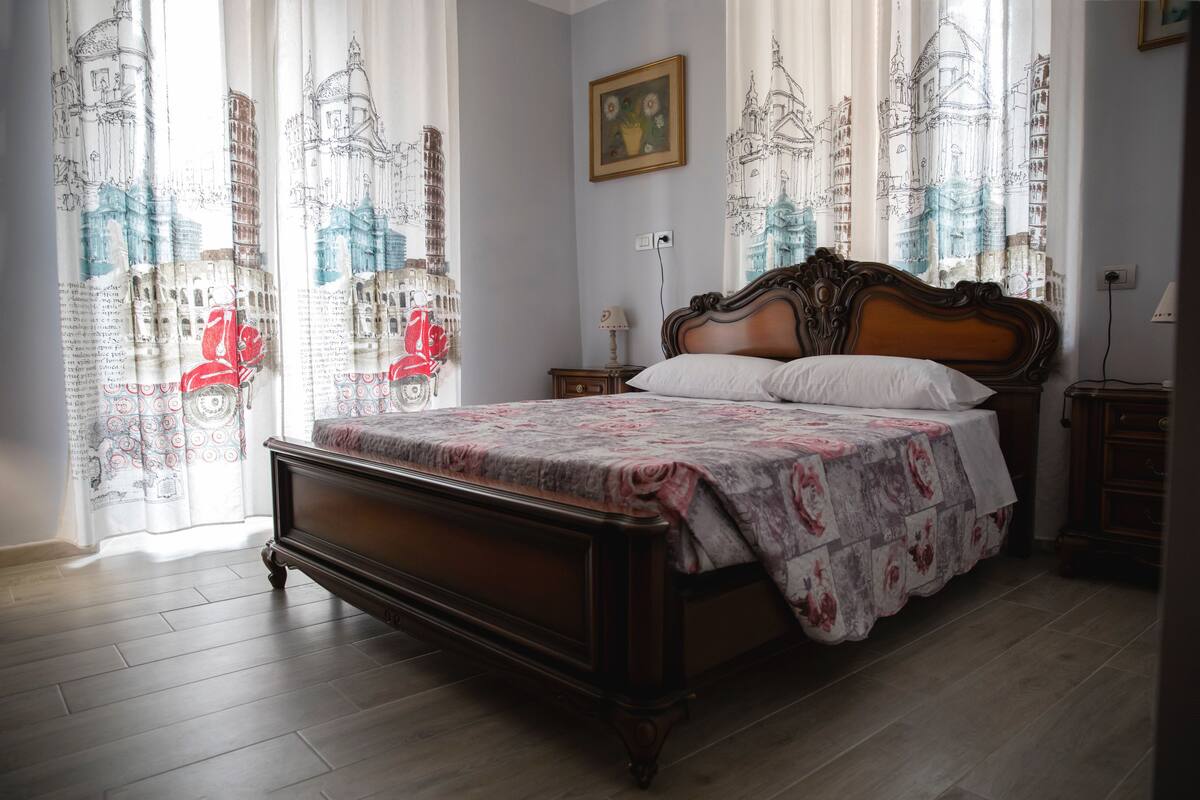 A bedroom with a wooden bed, floral bedding, and curtains with architectural drawings.