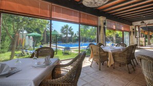 Un restaurant doté de mobilier en osier et de grandes fenêtres donnant sur une piscine et une végétation luxuriante.