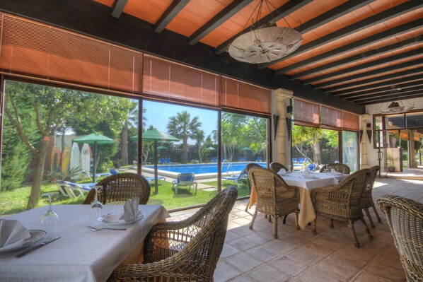 A restaurant with wicker furniture and large windows overlooking a swimming pool and lush greenery.