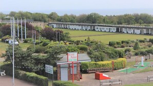 Un terrain de golf avec un green d’entraînement, un bâtiment en briques sur lequel on peut voir une enseigne, et une rangée de structures blanches en arrière-plan.