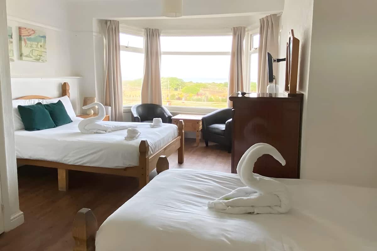 Ein Hotelzimmer mit zwei Betten, einem Holzkopfteil, einem Schreibtisch, einem Stuhl, einer Kommode und einem Fenster mit Vorhängen.