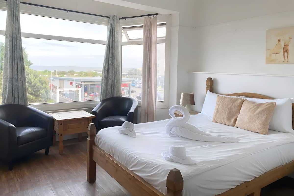 Ein Hotelzimmer mit einem großen Bett, zwei Sesseln, einem kleinen Tisch und Blick auf die Stadt durch große Fenster.