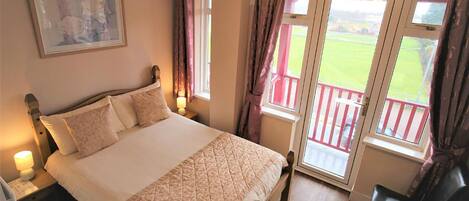 A bedroom with a large bed, a nightstand, a lamp, a window with curtains, and a view of a balcony.