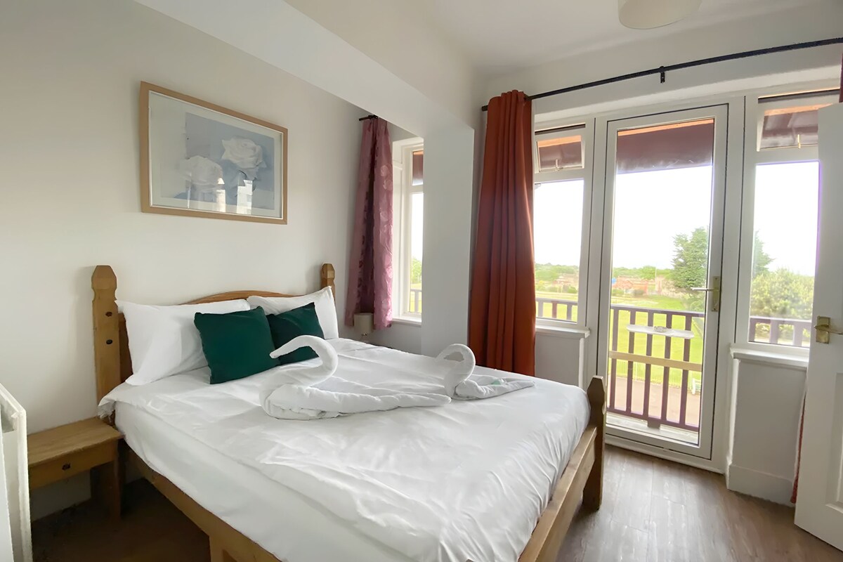 Ein ordentlich bezogenes Bett mit weißen Leinen, eine Schwan-Handtuch-Figur, ein Holzkopfstück, ein Nachttisch, ein gerahmtes Bild an der Wand und ein Balkon mit Blick auf Grünflächen.