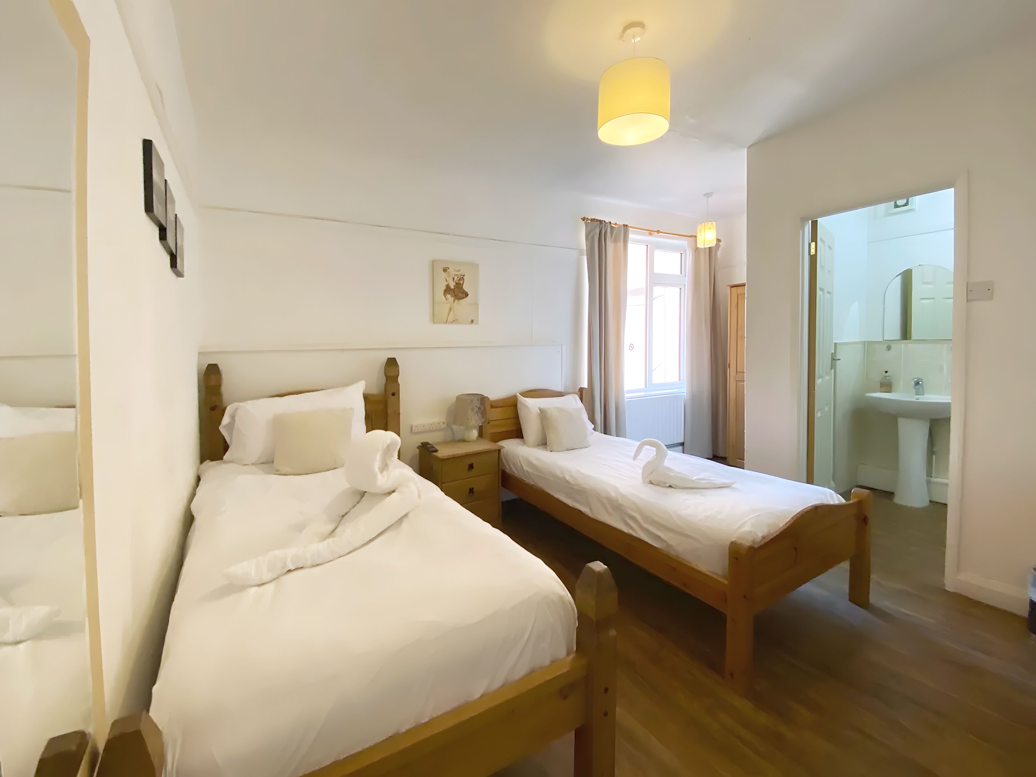 A clean, well-lit room with two wooden beds, a bedside table with a lamp, a painting on the wall, and a bathroom visible through an open door.