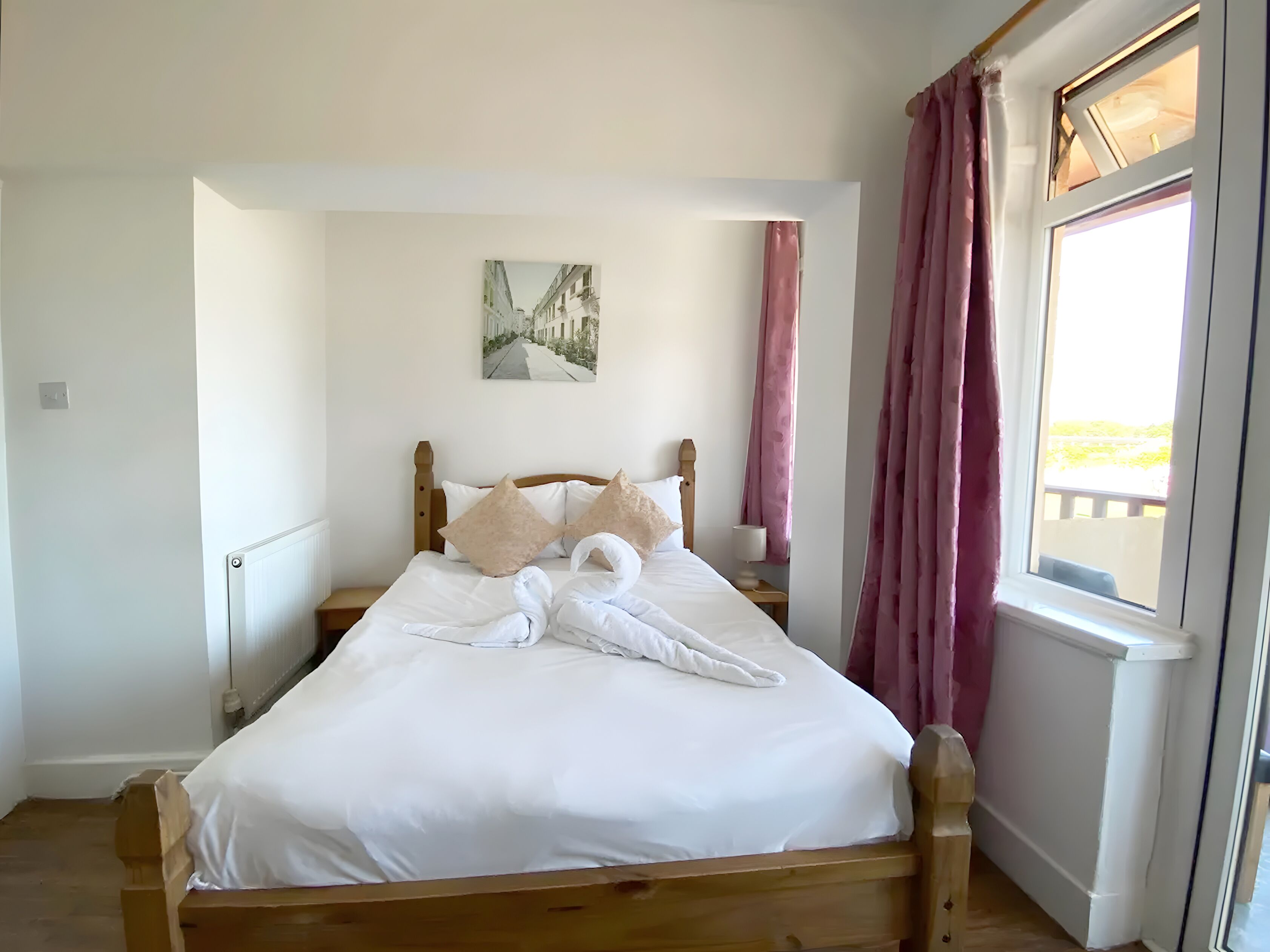 A neatly made bed with white linens and two pillows, a wooden headboard, a nightstand with a lamp, and a window with pink curtains.