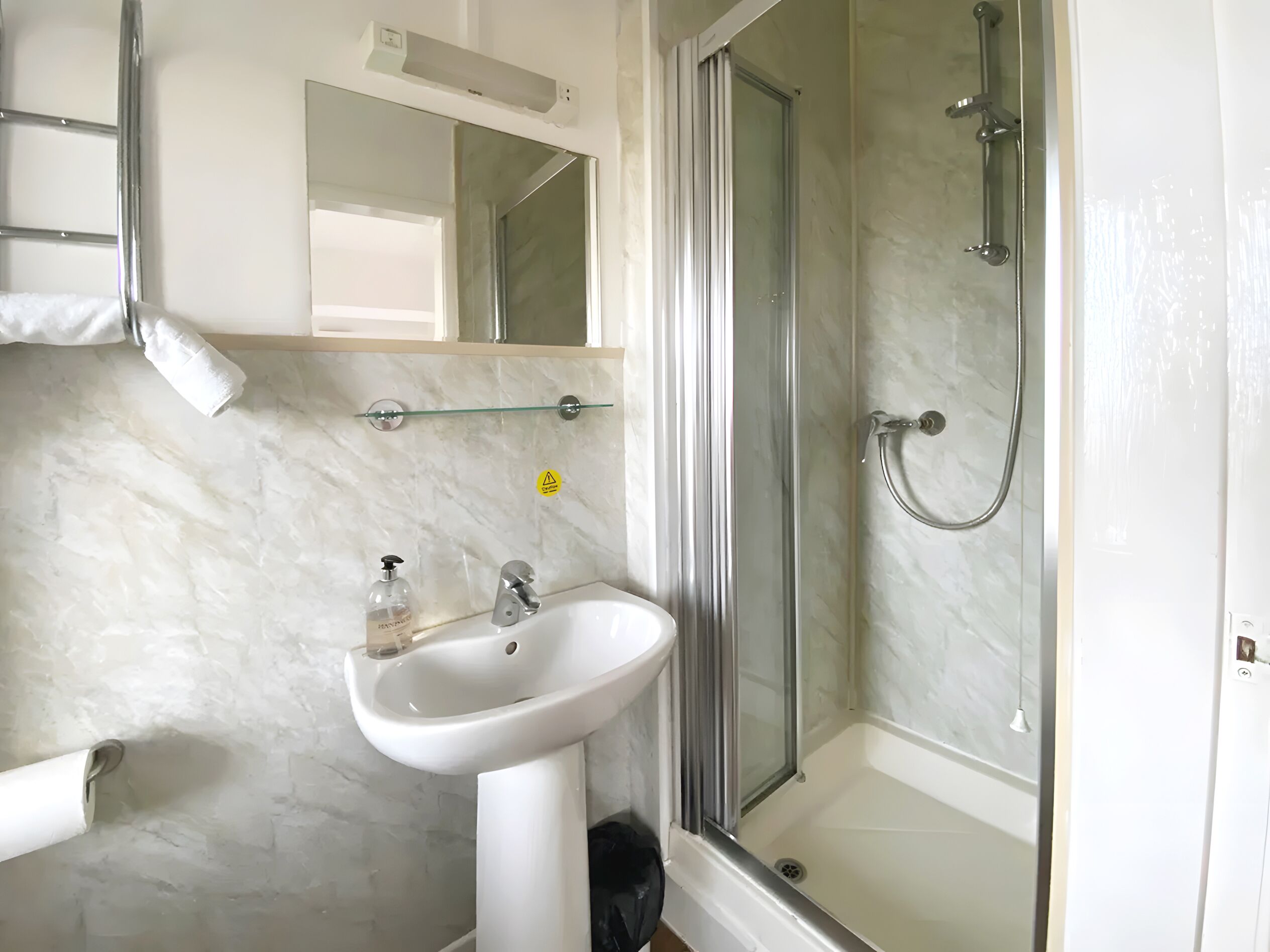 Een badkamer met een wit wastafeltje op voet, een douchecabine van glas en een spiegel boven het wastafeltje.