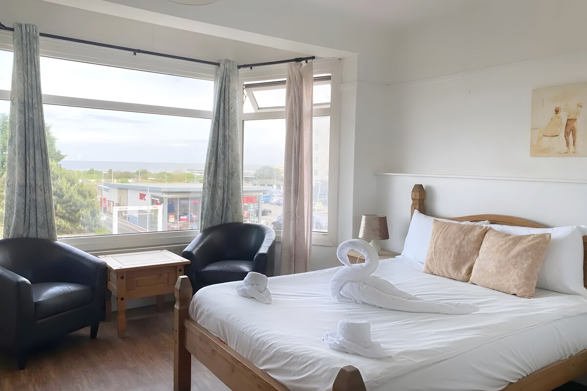 Une chambre d’hôtel avec un grand lit, deux fauteuils, une petite table et une vue sur la ville grâce à de grandes fenêtres.