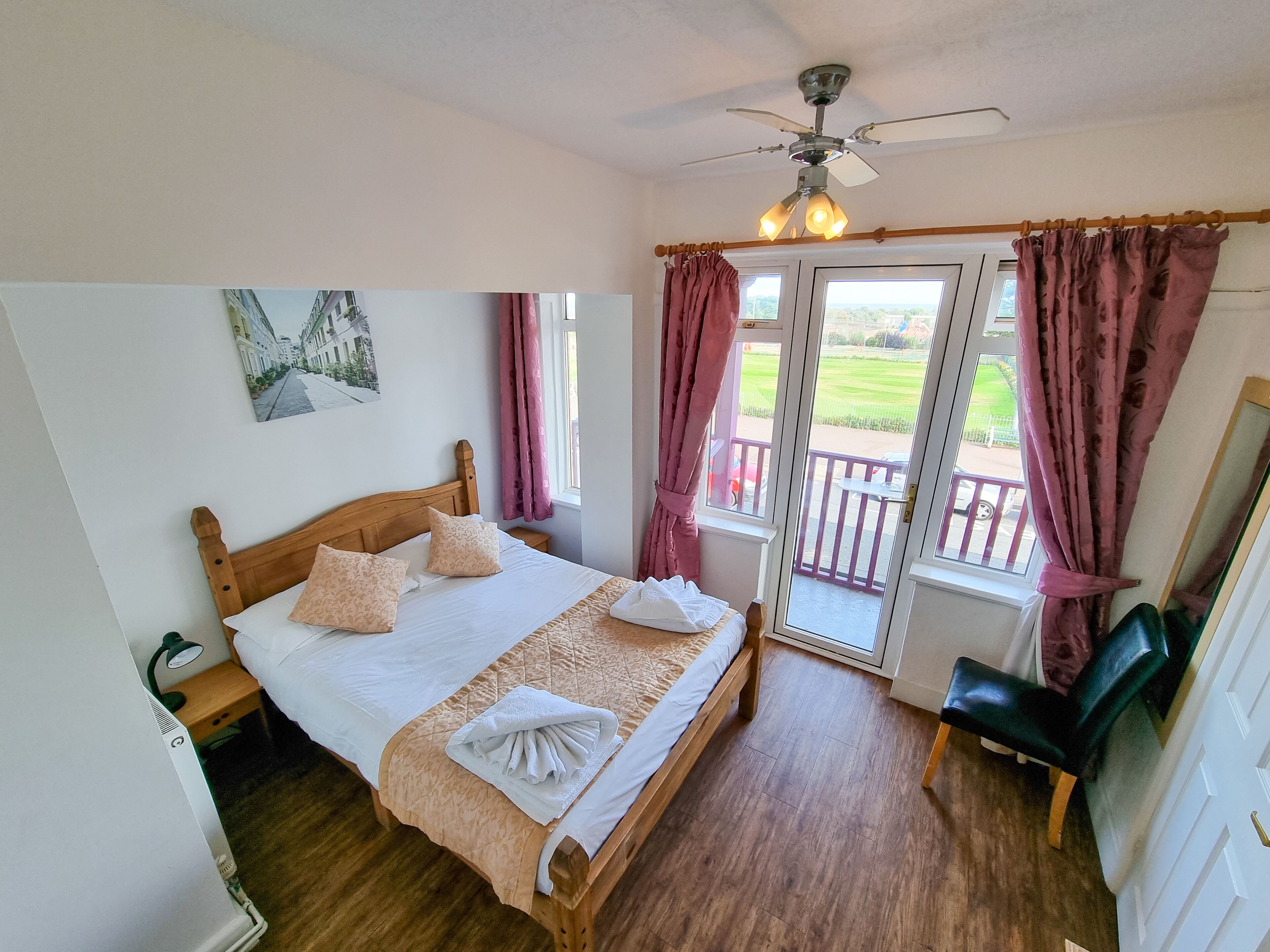 A neatly made bed with a wooden headboard, a bedside table with a lamp, a balcony with a view of greenery, and a ceiling fan.