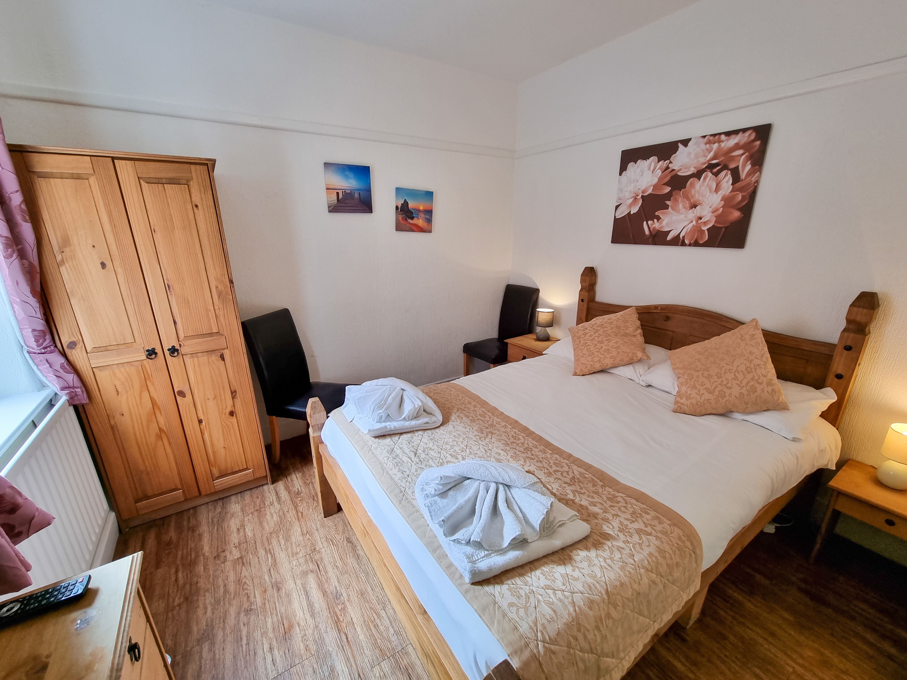 Une chambre bien rangée, avec une armoire en bois, un lit recouvert d’une literie à motifs, une chaise et deux tableaux accrochés au mur.