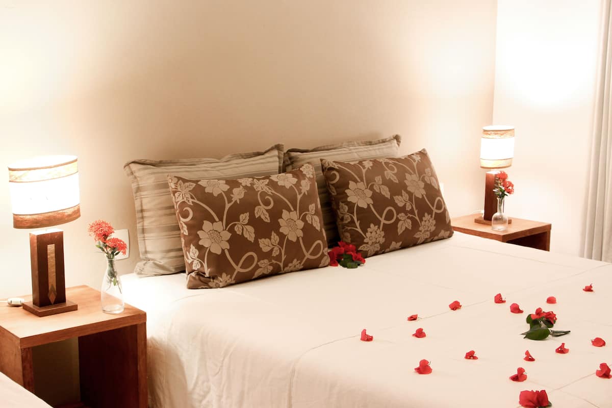 Cama bem arrumada com almofadas florais, um vaso com flores e pétalas de rosa sobre a cama.
