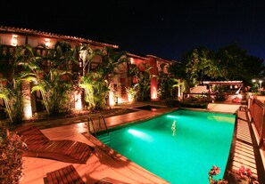 A swimming pool with a wooden deck and palm trees, surrounded by a building with red-tiled roofs and warm lighting. - Barcaça Pousada (Itacaré)