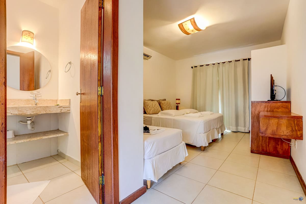 Quarto com duas camas, um guarda-roupa de madeira, banheiro com pia e espelho, e luminária no teto.