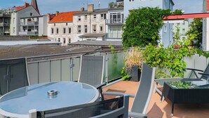 A rooftop terrace with a glass table and chairs, surrounded by buildings with balconies and a church spire in the background. - Haus Shiralee (Rostock)