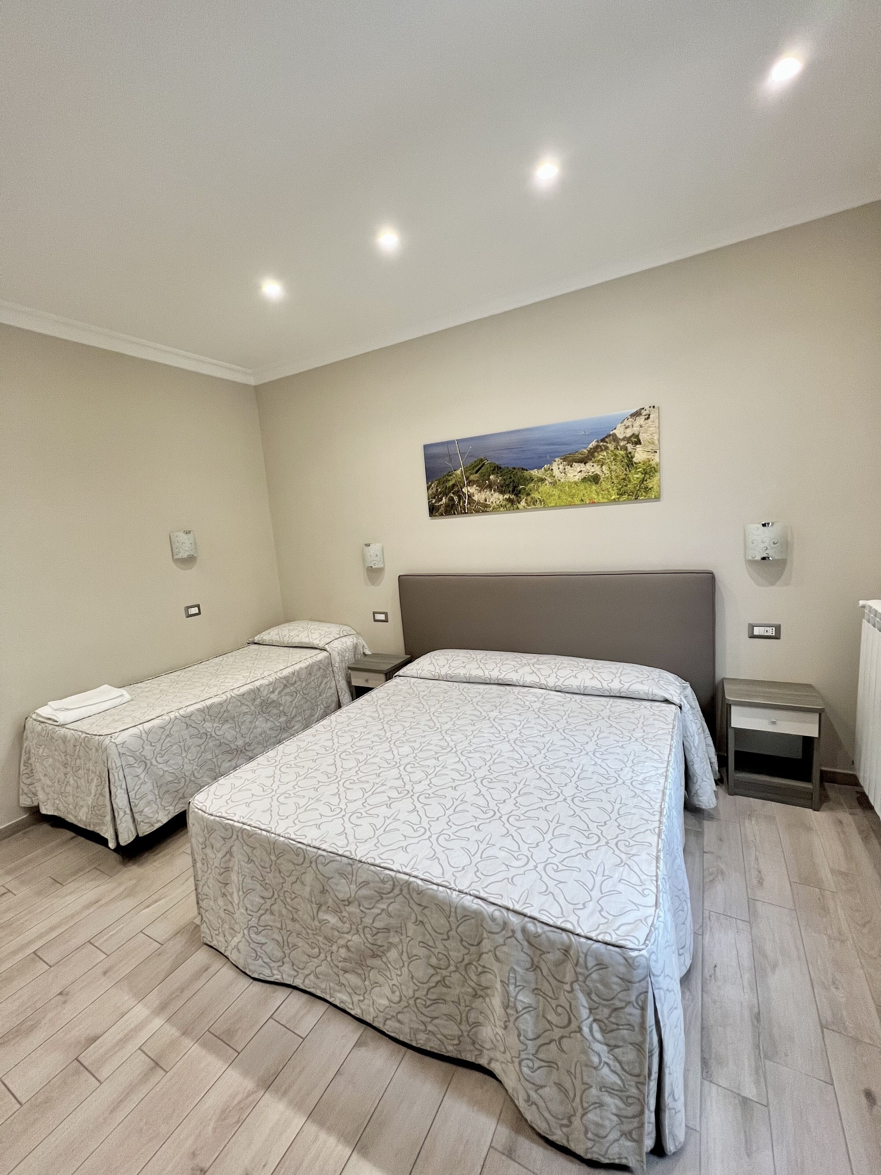 A bedroom with two single beds, a large bed, a painting of a coastal landscape, and a small bedside table with a lamp.