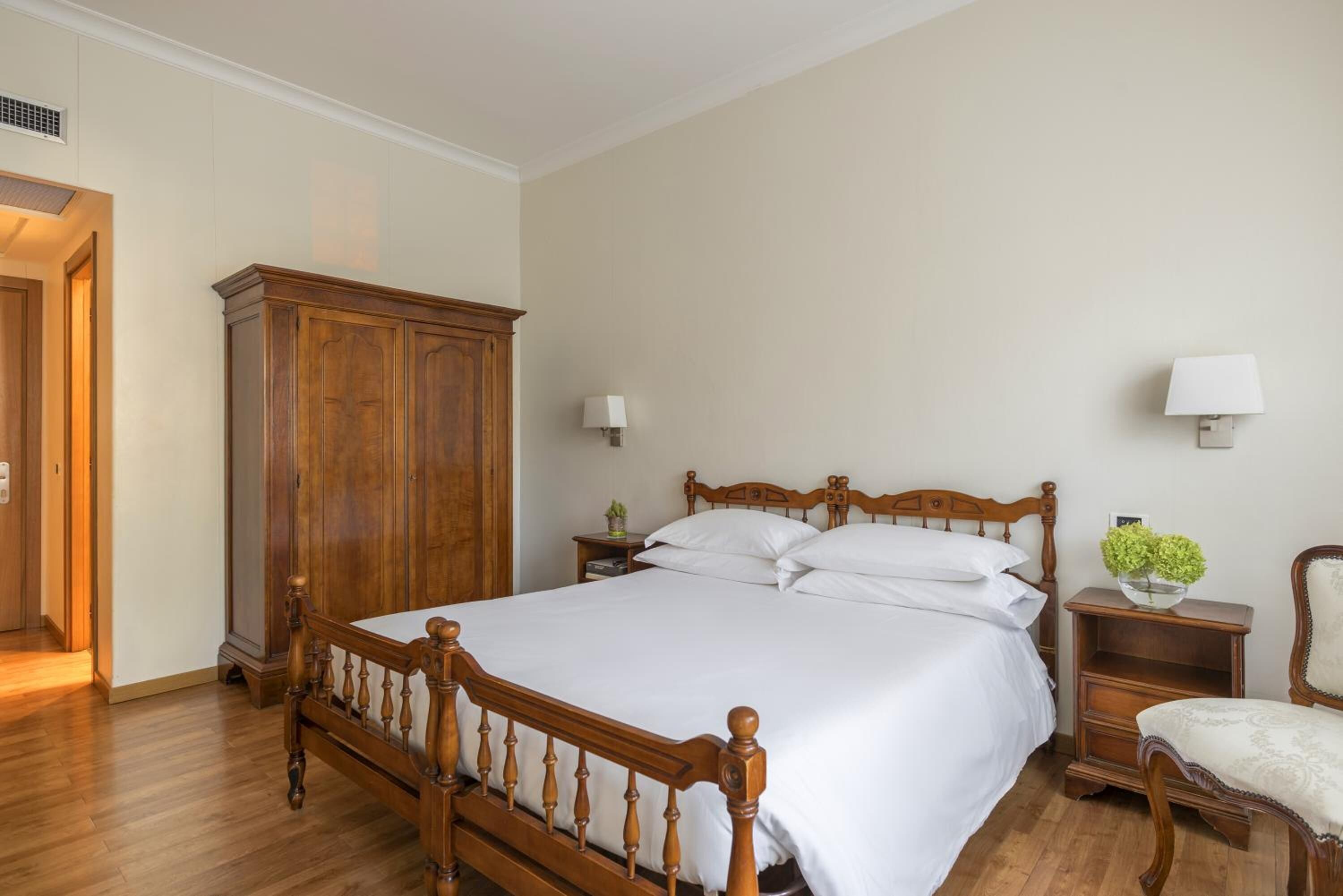 A bedroom with a wooden bed, a nightstand, a chair, a wardrobe, and two wall-mounted lamps.