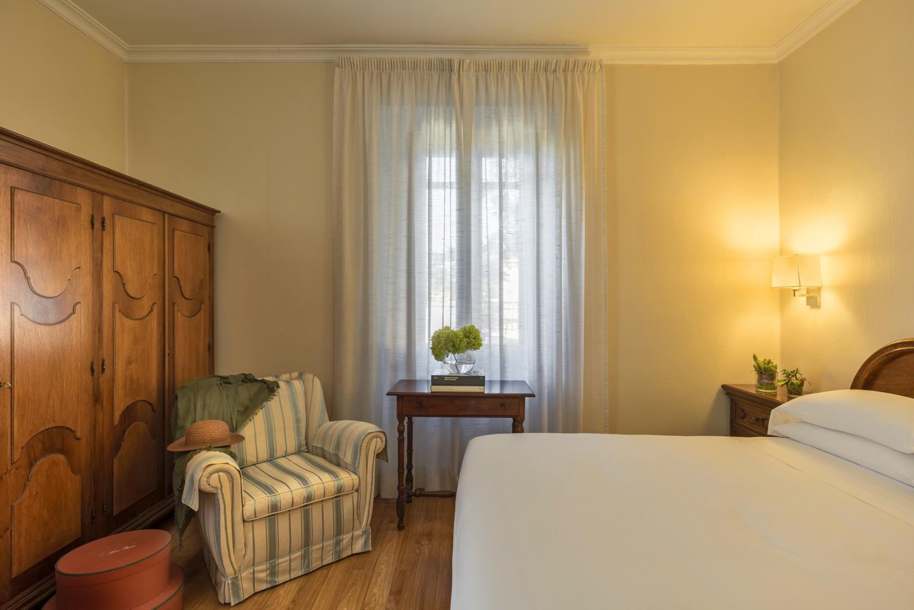 A hotel room with a bed, a wooden wardrobe, a striped armchair, a small table with a vase, and a nightstand with a lamp.