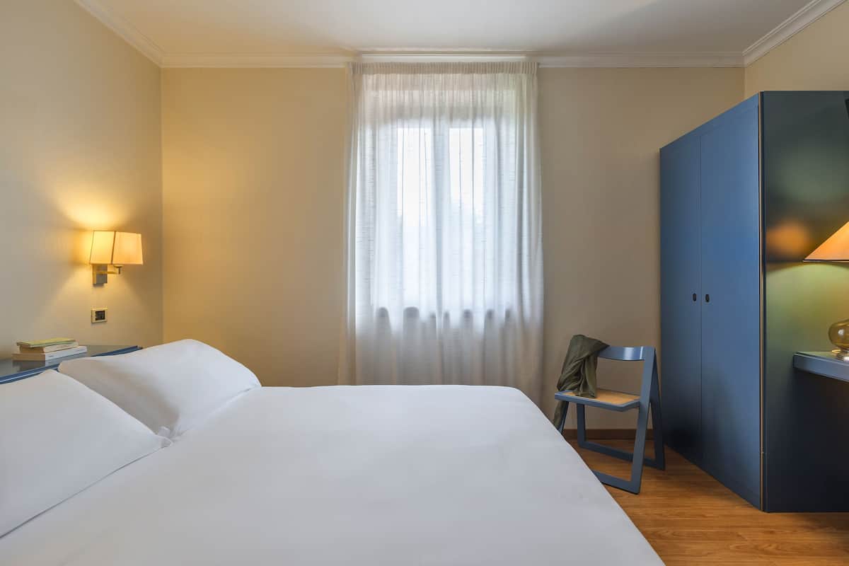 Ein Hotelzimmer mit Bett, Sessel, Kleiderschrank und einem fenster mit Vorhängen.