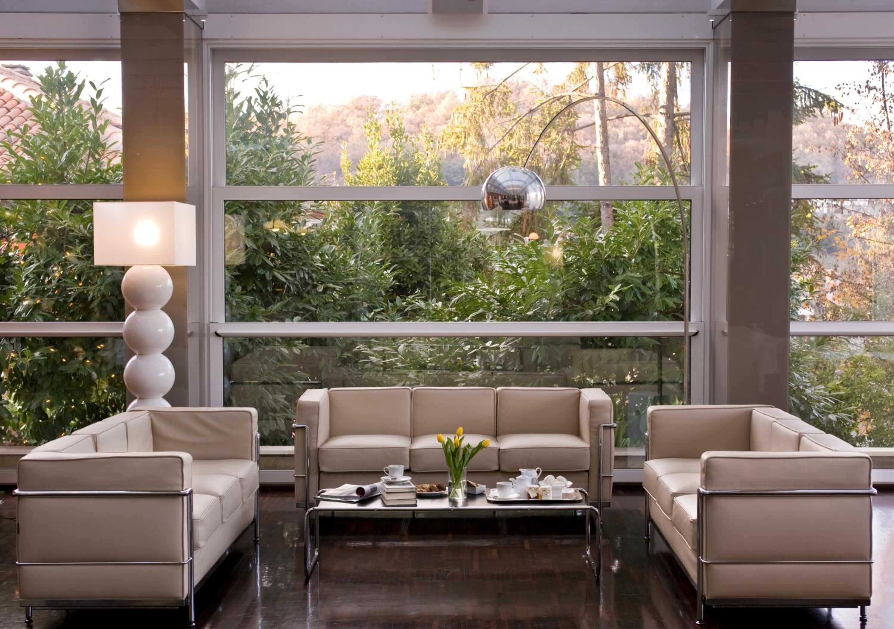 Un salon moderne avec un ensemble de canapés beige, une table basse agrémentée de livres et d’un vase de fleurs, et de grandes fenêtres offrant une vue sur la verdure.